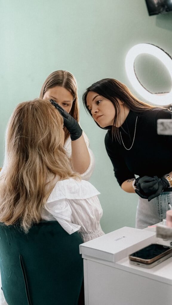 Two makeup artists apply cosmetics on a client in a beauty salon under ring light.